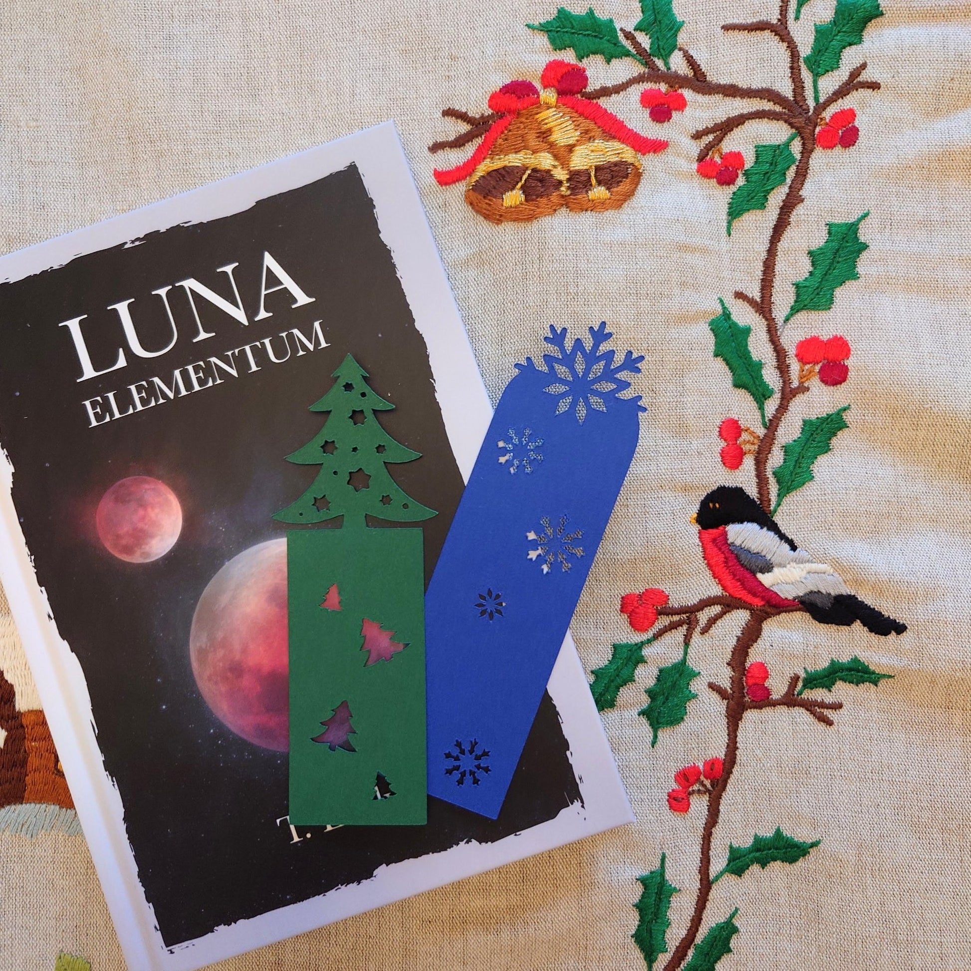 To bokmerker ligger oppå boken Luna elementum. En brodert juleduk i bakgrunnen.